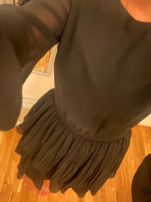 Svart volangklänning med lång ärm - Snygg svart kortklänning med volangkjol och transparenta långärmar. Klänningen har rund halsringning och ett flowy, luftigt tyg som ger en elegant look. Perfekt för dig som gillar stilrena och trendiga plagg med extra volym i kjolen. Fungerar både på vintern med boots och på sommaren med ballerina skor😉