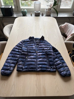 Blå camo pufferjacka från Polo Ralph Lauren - Snygg blå pufferjacka från Polo Ralph Lauren i camomönster. Jackan har dragkedja framtill, två fickor med dragkedja och en hög krage. Perfekt för kalla vinterdagar och ger en cool streetwear-look. Storleken är för dem som är 14-16 år
