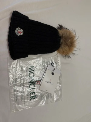 Svart Moncler mössa med pälsboll - Säljer en svart ribbstickad Moncler mössa med stor beige pälsboll på toppen och klassisk Moncler-logga framtill. Mössan är i tjockt, mjukt material och har en snygg, trendig look som passar perfekt till kalla dagar.