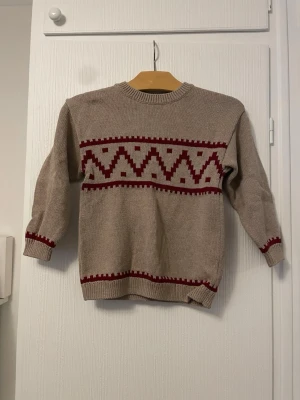 Beige stickad tröja med rött mönster H&M - Beige stickad tröja från H&M med röda zigzag- och rutmönster över bröstet och ärmsluten. Tröjan har rund halsringning och långa ärmar. Perfekt för kyligare dagar och enkel att matcha med jeans eller kjol.