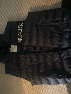 Svart dunväst från Moncler - Snygg svart dunväst från Moncler med stor vit logga broderad på bröstet. Västen har en hög krage, dragkedja framtill och en ficka med dragkedja. Perfekt för lager-på-lager och ger en clean streetwear-look.