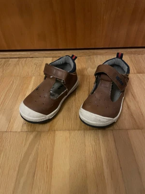 Bruna barnskor med kardborre - Säljer ett par bruna barnskor från Mino med kardborreband och vita sulor. Skorna har öppen design framtill, mörkblå detaljer och en liten rödblå flik bak. Perfekta för lek och vardag.