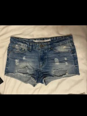 Blå slitna jeansshorts - Säljer ett par blå jeansshorts med slitna detaljer och råa kanter. Shortsen har fem fickor, bälteshällor och knappgylf. Perfekta för sommaren och ger en avslappnad vibe. Det står M men skulle mer säga S i storlek.