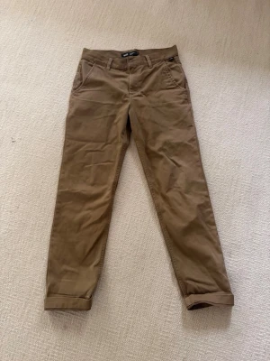 Beiga chinos med raka ben VANS stl29/18 - Snygga beiga chinos med raka ben och klassisk passform. Byxorna har sidofickor fram och två bakfickor med knapp. Tillverkade i ett mjukt bomullsmaterial som känns skönt mot huden. Litet märke vid vänster ficka, men det är inte hål.