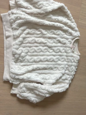 Vit fluffig tröja med strukturmönster - Supermjuk och fluffig vit tröja med vågigt strukturmönster över hela plagget. Tröjan har rund halsringning, långa ärmar och breda ribbade muddar vid ärmslut och nederkant. Perfekt för dig som gillar mysiga och trendiga plagg.
