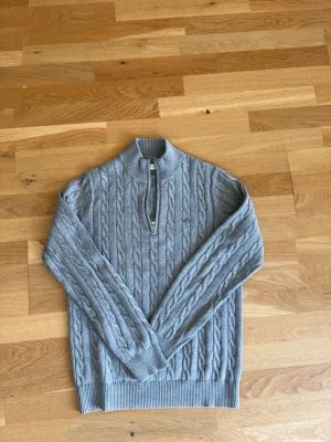 Ljusgrå kabelstickad tröja med halvzip från gant - Snygg ljusgrå kabelstickad tröja med halv dragkedja och hög krage. Tröjan har ribbade muddar vid ärmslut och nederkant, samt ett diskret broderat märke på bröstet. Perfekt för lager på lager och en clean look.