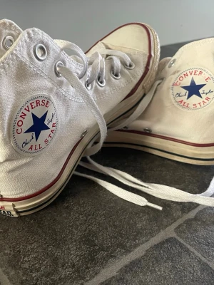 Vita Converse stl 37,5 - Klassiska vita Converse Chuck Taylor All Star high-tops med canvasmaterial, vita snören och gummisula. Skorna har den ikoniska blå stjärnloggan på sidan och röda samt svarta detaljer längs sulan. Perfekta för en avslappnad och cool stil.