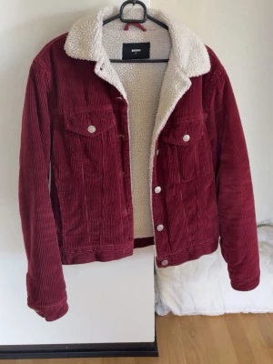 Vinröd teddyjacka från Bik Bok - Vinröd teddyjacka från Bik Bok i manchestertyg med vit, fluffig teddyfoder på insidan och kragen. Jackan har två bröstfickor med knappar och silverfärgade detaljer. Perfekt för att hålla sig varm och snygg under kyliga dagar.