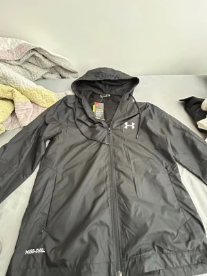 Svart vindjacka från Under Armour - Snygg svart vindjacka från Under Armour med huva, dragkedja framtill och vita logodetaljer. Jackan har meshfoder för extra ventilation och är tillverkad i lätt, tekniskt material. Perfekt för aktiva dagar utomhus. Under armour helt ny med prislapp behöver sälja på grund av pengarna 