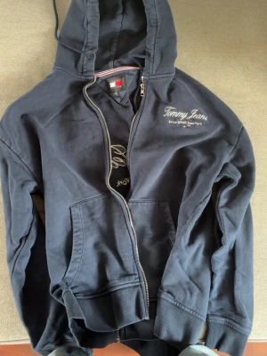 Marinblå hoodie från Tommy Jeans - Säljer en marinblå hoodie från Tommy Jeans med dragkedja och broderad logga både fram och bak. Klassisk huva med snörning och kängurufickor. Tillverkad i mjukt bomullsmaterial, perfekt för en avslappnad och stilren look.