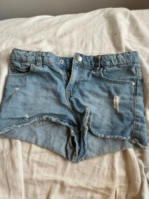 Ljusa jeansshorts med slitningar - Säljer ett par ljusblå jeansshorts med rå kant och slitna detaljer. Klassisk femficksmodell med bälteshällor och knappgylf. Perfekta för sommaren och ger en avslappnad vibe till din outfit.