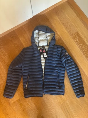 Mörkblå colmar dunjacka - Snygg mörkblå dunjacka med quiltad design och huva. Jackan har dragkedja framtill, två sidofickor med dragkedja och en ljus insida. Perfekt för kalla  höstdagar och riktigt skön att bära. Jackan är i storlek M