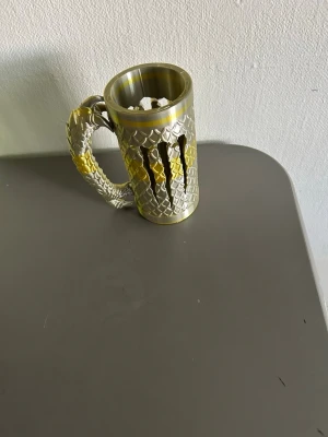 Monster hållare  - Unik mugg i metall med drakfjällsmönster och guldfärgade detaljer. Handtaget är format som en draksvans och muggen har ett fantasyinspirerat utseende. Perfekt för dig som gillar fantasy eller vill ha något speciellt till drycken.