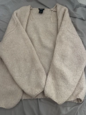Beige stickad kofta från Monki - Mysig beige stickad kofta från lindex med öppen front och avslappnad passform. Koftan har långa ärmar och är gjord i ett mjukt, fluffigt material som känns skönt mot huden. Perfekt att slänga över en t-shirt eller topp för extra värme.