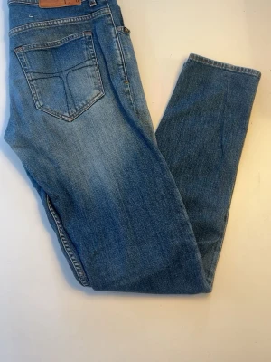Blå raka jeans i klassisk modell - Ett par blå jeans med rak passform och fem fickor. Jeansen har en klassisk ljus tvätt och subtila slitningar framtill. Tillverkade i denim med normal midja och bälteshällor. Perfekta för en avslappnad och tidlös look. Tiger Of Sweden närket