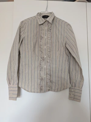 Randig skjorta med volangdetalj - Snygg skjorta från Woman's Taylor med smala ränder i beige, grått och ljusgrönt. Skjortan har volangdetaljer längs knappraden och klassisk krage. Perfekt för dig som gillar en stilren men ändå lekfull look.