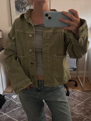 Olivgrön jeansjacka med fickor - Snygg olivgrön jeansjacka med två stora framfickor och klassisk krage. Jackan har silverfärgade knappar framtill och en avslappnad passform som ger en cool och trendig look. Perfekt att slänga över en t-shirt för en casual stil.