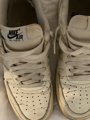 Nike Air Force 1 vita sneakers - Klassiska vita Nike Air Force 1 sneakers med lågt skaft. Skorna har perforerad tå, vita skosnören och den ikoniska svarta Nike-loggan på sidan. Ovandelen är i läder och sulan är vit med en lätt gulnad kant. Perfekta för en clean och tidlös streetstil.