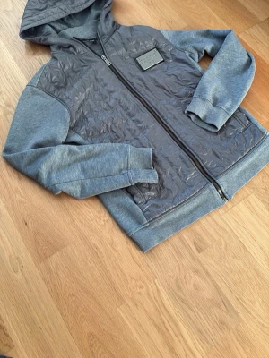  Dolce & Gabbana - Snygg grå hoodie från Dolce & Gabbana Men's Collection med quiltad front och huva. Jackan har dragkedja och ribbade muddar. Materialmix med polyester på framsidan och mjuk bomull på ärmar och baksida. Perfekt för dig som gillar exklusiva streetwear-vibes. 