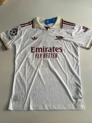 Helt ny! Arsenal 25/26 tredje EZE  - Snygg vit Arsenal matchtröja från Adidas med vinröda detaljer och trycket EZE 10 på ryggen. Tröjan har Champions League-märke på ärmen, klassisk krage och diskret mönster i tyget. Tillverkad i lätt funktionsmaterial, perfekt för fotboll eller träning.
