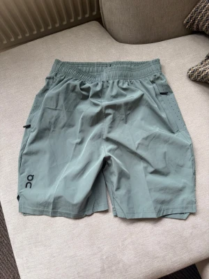 On Running Shorts – grågröna, storlek M - Säljer ett par stilrena träningsshorts från On i grågrön färg. Lätt och följsam sportmodell med elastisk midja och praktiska fickor med dragkedja.  Storlek M. Shortsen är i fint bruksskick och passar perfekt för löpning, gym, träning eller vardag.  