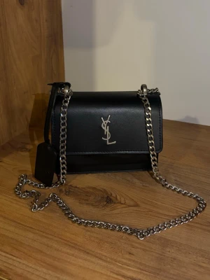 Svart YSL axelväska i läder - Snygg svart axelväska i slätt läder med silverfärgad kedja och logga framtill. Kompakt och stilren design med klassiska detaljer. Perfekt för dig som vill ha en trendig och lyxig accessoar till din outfit.