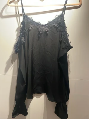 Svart omlottopp med spetsdetaljer - Snygg svart omlottopp med tunna axelband och dekorativa spetsdetaljer längs ringningen. Toppen har offshoulder-långa ärmar och är tillverkad i ett glansigt material som ger en lyxig känsla. Perfekt för dig som vill ha en trendig och elegant look.