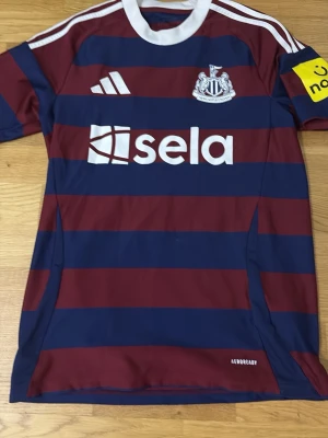 Randig Newcastle matchtröja - Snygg Newcastle matchtröja från Adidas med breda vinröda och marinblå ränder. Tröjan har vita detaljer, klubbmärke på bröstet och sponsorlogga framtill. Tillverkad i lätt och ventilerande material, perfekt för fotboll eller träning.