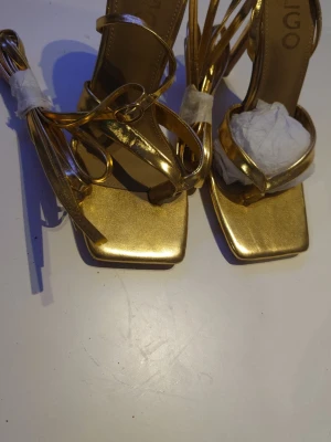 Guldiga sandaletter med snörning - Säljer ett par glansiga guldiga sandaletter med fyrkantig tå och tunna remmar som snörs runt ankeln. Sandalerna har en elegant design och klack, perfekta för att sticka ut. Materialet är syntetiskt med metallic-finish.