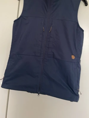 Mörkblå väst från Fjällräven - Stilren mörkblå väst från Fjällräven med dragkedja framtill och två fickor med dragkedja. Västen har det klassiska Fjällräven-logotypen i läder på bröstet och är tillverkad i slitstarkt material. Perfekt för lager-på-lager och outdoor.