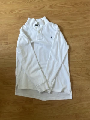 Vit långärmad raulph lauren - Klassisk vit långärmad henley med två knappar i halsen och liten broderad logga på bröstet. Skjortan har en rak passform och är tillverkad i mjuk bomull, perfekt för en clean och enkel stil.