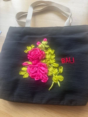 Svart tottebag med broderade rosor - En svart shoppingväska/ totte bag med naturvita handtag och broderade rosa rosor samt gröna blad på framsidan. Texten 'BALI' är broderad i rött. Väskan har en mjuk struktur och är perfekt för att bära med sig det viktigaste.