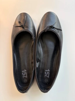 Svarta ballerinaskor med rosett - Stilrena svarta ballerinaskor med liten rosett framtill. Skorna har rundad tå och är tillverkade i ett slätt, syntetiskt material. Yttersulan är mönstrad för bättre grepp. Perfekta för en enkel och klassisk look.