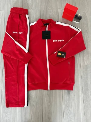 Rött tracksuit set Palm Angels - Snyggt rött tracksuit set från Palm Angels med vita detaljer längs ärmar och ben. Setet består av en zip-jacka och matchande byxor, båda med logga på bröst och lår. Tillverkat i mjukt, glansigt material som ger en sportig och trendig look.