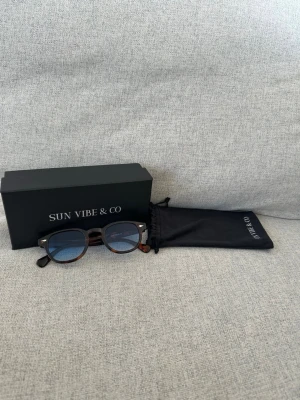 Solglasögon från sunvibe - Snygga runda solglasögon från Sun Vibe & Co med brunmelerad båge och blåtonade glas. Kommer med svart fodral och originalask. Perfekta för dig som vill ha en stilren accessoar med en modern touch.