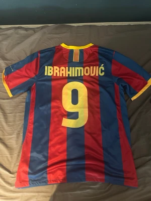 Barcelona Ibrahimović matchtröja - Säljer en klassisk FC Barcelona matchtröja med Ibrahimović och nummer 9 på ryggen. Tröjan har blå och röda vertikala ränder, gula detaljer vid krage och ärmslut samt klubbmärke och Nike-logga på bröstet. Tillverkad i lätt och ventilerande material, perfekt för fotboll.