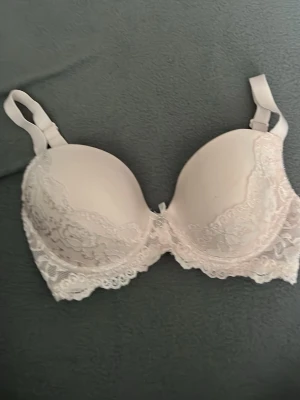 Ljusrosa push-up bh med spets - Söt ljusrosa bh med vadderade kupor och push-up effekt. Dekorerad med spetsdetaljer och en liten rosett med metallhänge framtill. Justerbara axelband och mjukt material för extra komfort.