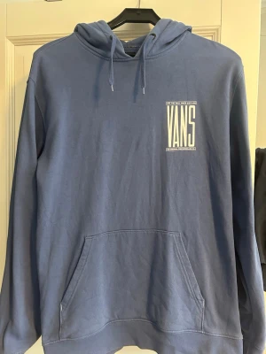 Blå hoodie från Vans - Säljer en blå hoodie från Vans med stor vit logga på ryggen och mindre logga på bröstet. Klassisk känguruficka framtill och justerbar huva med dragsko. Perfekt för dig som gillar streetwear och vill ha en skön och stilren look. Hoodien har bara användts ett par gånger och är i perfekt skick.
