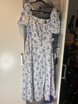 Blommig vit och blå klänning - Söt lång klänning från Shein i vitt med blått blommönster. Klänningen har puffärmar, smockad överdel och knytning framtill. Tillverkad i ett lätt och luftigt material, perfekt för sommarens alla tillfällen. Använd 1 gång
