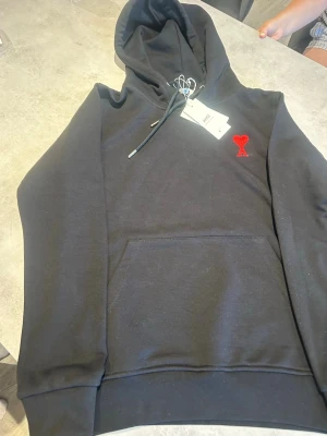 Svart hoodie från Ami med röd logga - Säljer en svart hoodie från Ami med den ikoniska röda hjärtloggan broderad på bröstet. Hoodien har dragsko i huvan och en stor magficka. Tillverkad i mjuk bomullsblandning som känns skön mot huden. Perfekt för en chill och stilren look.