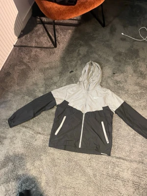 Nike tvåfärgad vindjacka med huva - Snygg vindjacka från Nike i svart och ljusgrått med dragkedja framtill och två fickor med dragkedja. Jackan har huva med dragsko och en vit Nike-logga på bröstet. Perfekt för blåsiga dagar och sportiga outfits.