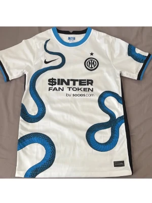 Vit Inter Milan matchtröja med orm - Säljer en vit Inter Milan matchtröja från Nike med blå orm-mönster och svarta detaljer. Tröjan har klubbmärke och Nike-logga på bröstet samt sponsortryck på framsidan. Rund halsringning med blå kant och korta ärmar. Perfekt för fotbollsfans!
