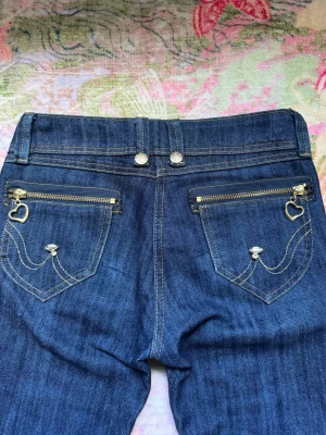 Snygga Lågmidjade bootcut jeans med detaljer - Fina mörkblå bootcut jeans med låg midja från Rebers Jeans😍De har dekorativa guldknappar och kontrastsömmar. Även dragkedja med hjärta och glittriga stenar på bakfickorna🩷Klassisk femficksmodell med låg midja och lätt slitna detaljer framtill! Perfekta för en avslappnad och trendig look🤍Passar så bra till alla plagg och skapar en unik outfit som drar uppmärksamhet🙌Storlek XXS, använda några gånger men utan defekter. Mått cm: midja 33, gren 19, innerben 83❤️Älskar dessa jeans med coola Y2k/2000s.