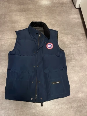 Mörkblå dunväst från Canada Goose - Snygg mörkblå dunväst från Canada Goose med hög krage, två stora framfickor och klassisk logotyp på bröstet. Västen har dragkedja och tryckknappar framtill samt mjukt innerfoder. Perfekt för lager-på-lager och håller dig varm under kyliga dagar.