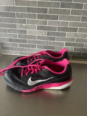 Svarta och rosa Nike sneakers - Snygga sneakers från Nike i svart med rosa detaljer och snören. Skorna har en vit sula och den klassiska Nike-loggan i silver på sidan. Ovandelen är i mesh och syntetmaterial för bra andningsförmåga.