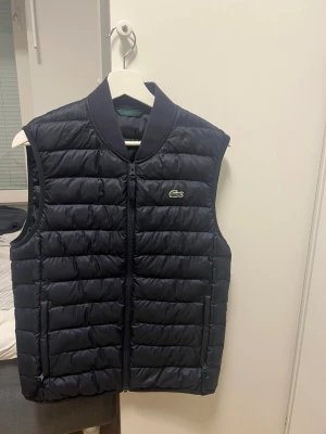Mörkblå dunväst från Lacoste - Snygg mörkblå dunväst från Lacoste med klassisk quiltad design och dragkedja framtill. Västen har ståkrage och den ikoniska krokodilloggan på bröstet. Perfekt för lager-på-lager och håller dig varm under kyliga dagar.