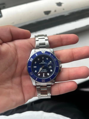 R0X Submariner blå och silver klocka - Stilren RX Submariner med blå urtavla och vridbar blå bezel. Klockan har en robust länk i silverfärgat stål och tydliga indexmarkeringar. Datumfönster vid klockan tre och klassisk RX-logga på spännet. Perfekt accessoar för dig som gillar exklusiv stil.