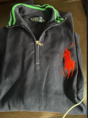 Mörkblå tröja från Polo Ralph Lauren - Mörkblå tröja med halv dragkedja från Polo Ralph Lauren. Stor röd logga på bröstet och krage med gröna ränder. Perfekt för en sportig och avslappnad stil. Mjuk och bekväm, passar till många tillfällen.