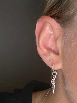 Silvrigt örhänge med spike charm - Säljer ett stilrent silvrigt örhänge med en liten ring och en hängande spetsig charm. Ovanför spetsen sitter en klar sten som ger extra bling. Perfekt för dig som gillar minimalistiska men ändå unika smycken.