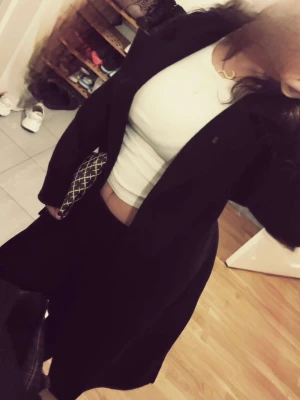 Svart lång kappa med klassisk krage - Snygg svart lång kappa med klassisk krage och rak passform. Kappan har en stilren look och passar perfekt till lager-på-lager outfits. Tillverkad i ett tjockare tyg som ger en elegant siluett. Enkelknäppt framtill och utan synliga detaljer.
